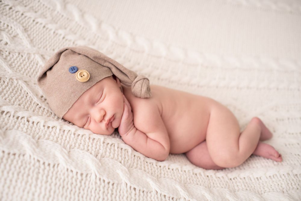 Sleeping newborn wearing brown knit hat on white textured blanket during photo session.