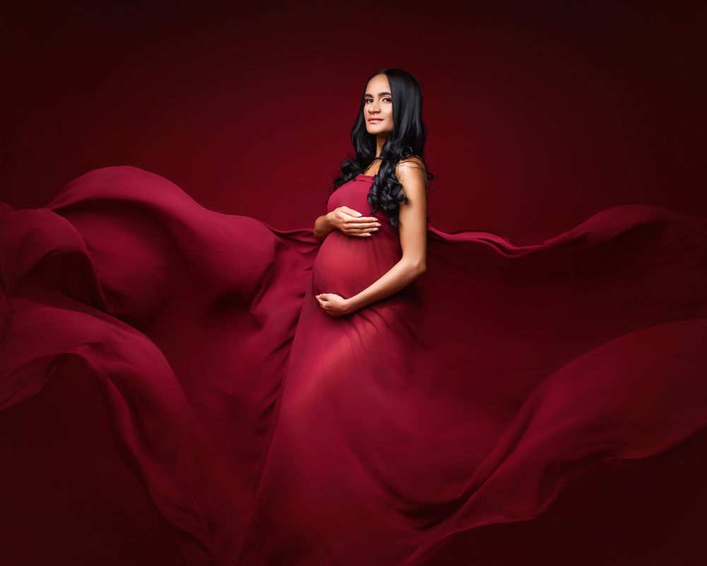 Artistic maternity portrait with flowing red fabric against dark red background.