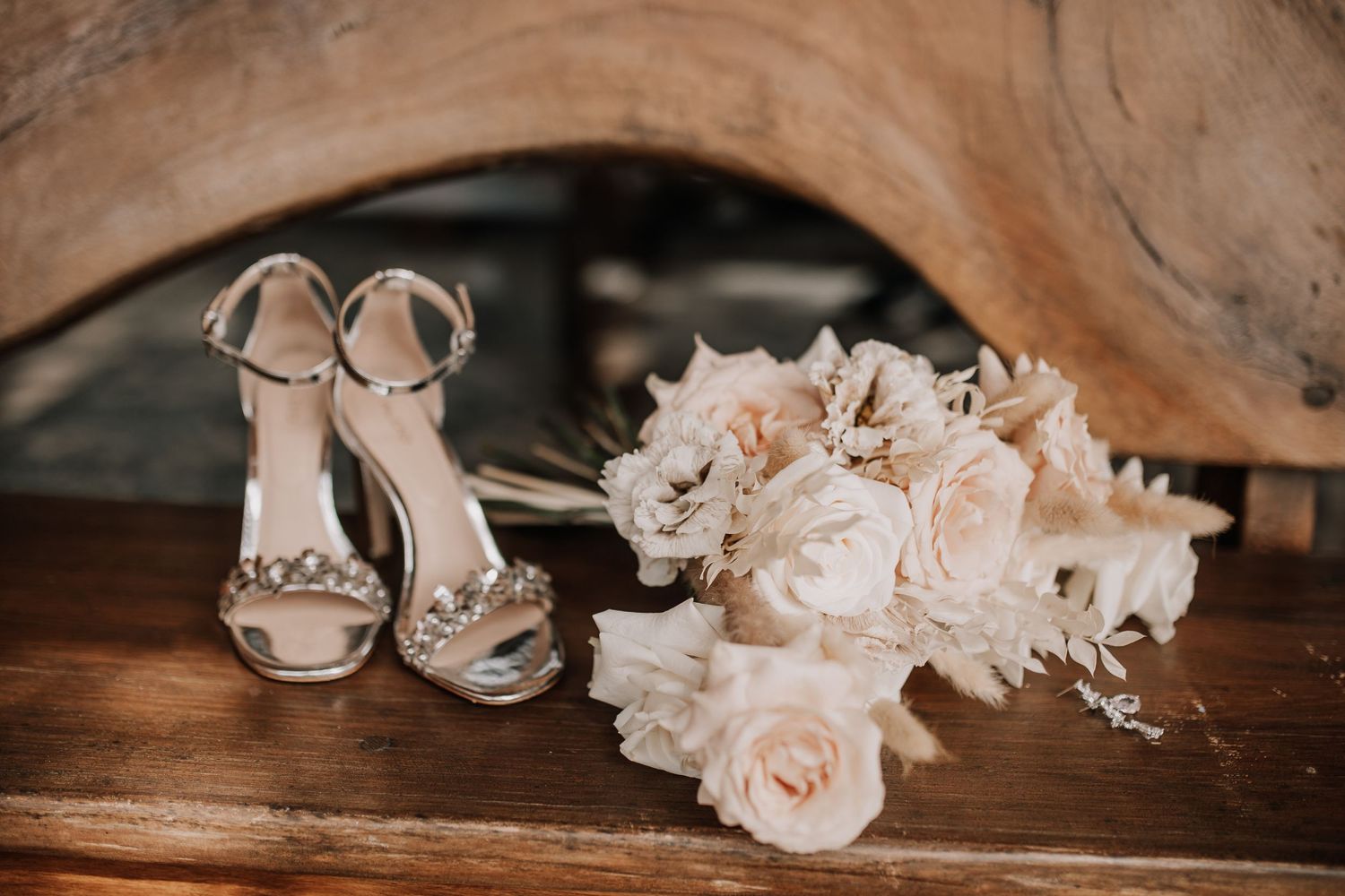 Silver bridal heels and white rose bouquet displayed on rustic wooden surface.