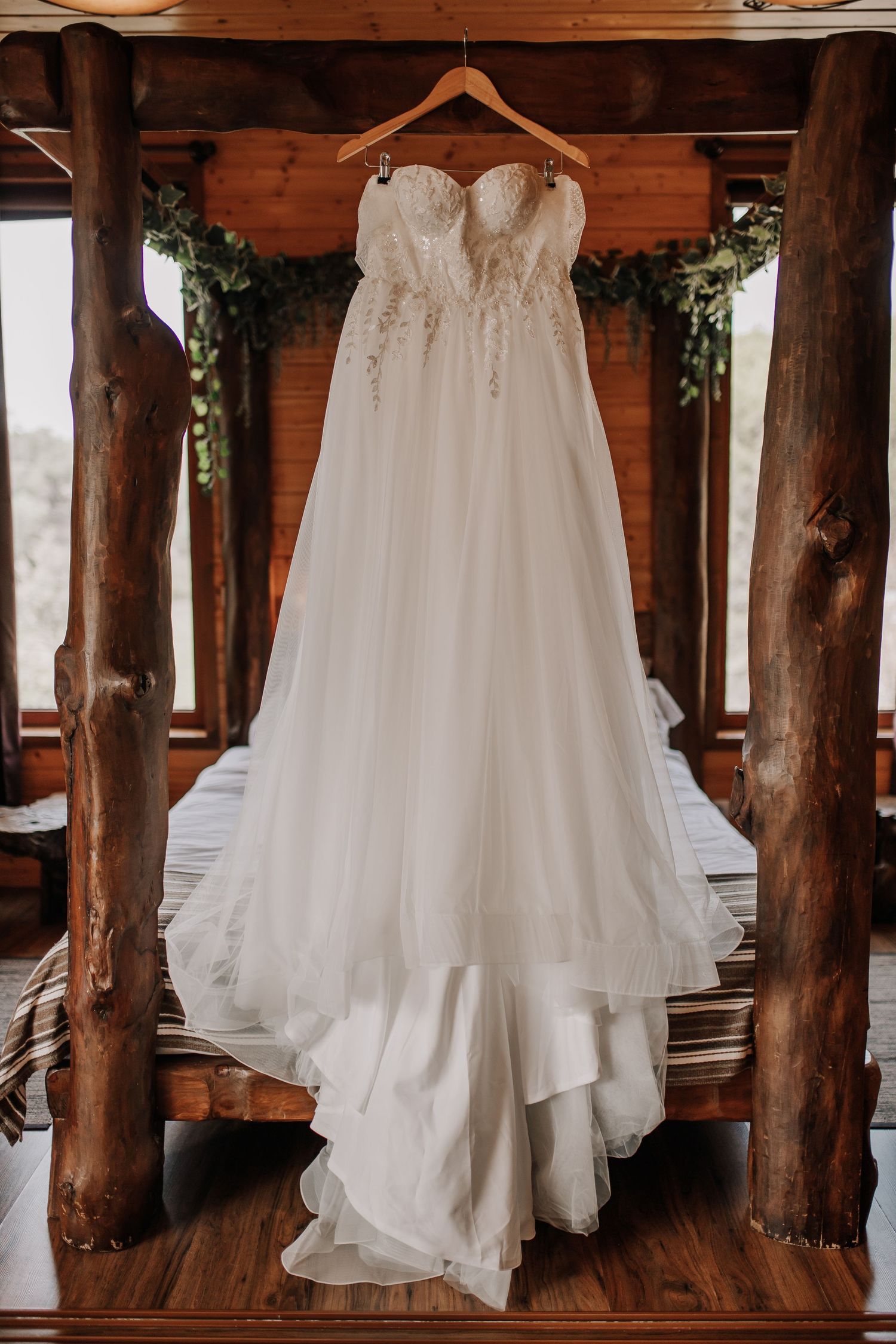 Elegant strapless lace wedding gown hanging on wooden hanger in rustic log cabin bridal suite.