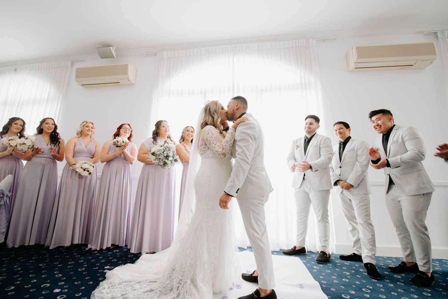 Wedding ceremony with bridesmaids in purple and groomsmen in white suits watching the couple's first kiss.