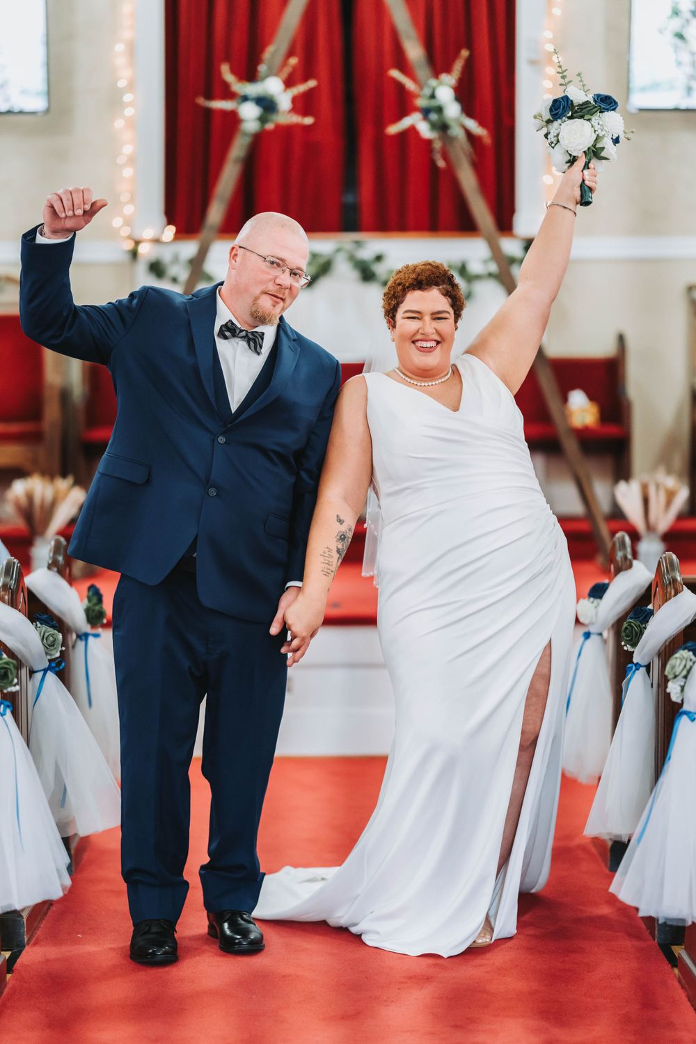 A newlywed couple celebrates with raised arms while walking down the red-carpeted church aisle.