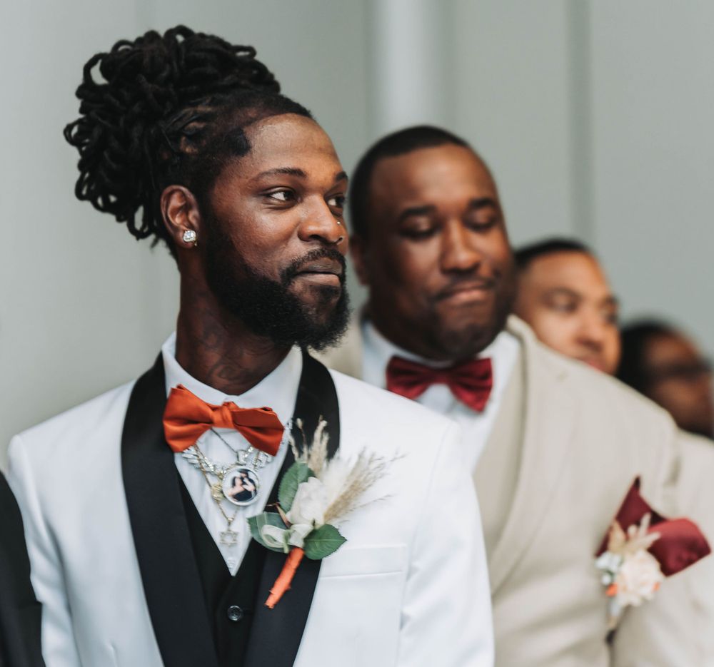 Groomsmen in white tuxedos and red bow ties stand at attention during wedding ceremony.