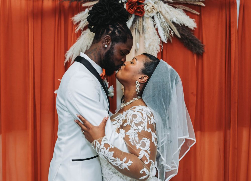 A couple shares a romantic kiss against an orange backdrop during their wedding ceremony.