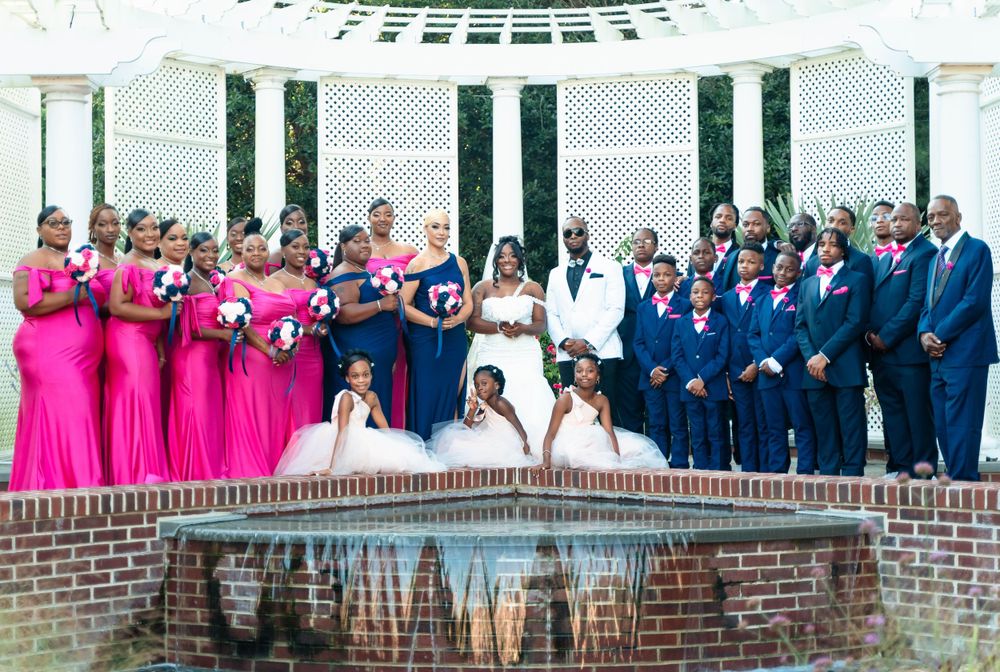 Large wedding party in bright pink and navy blue formal attire posing by fountain.