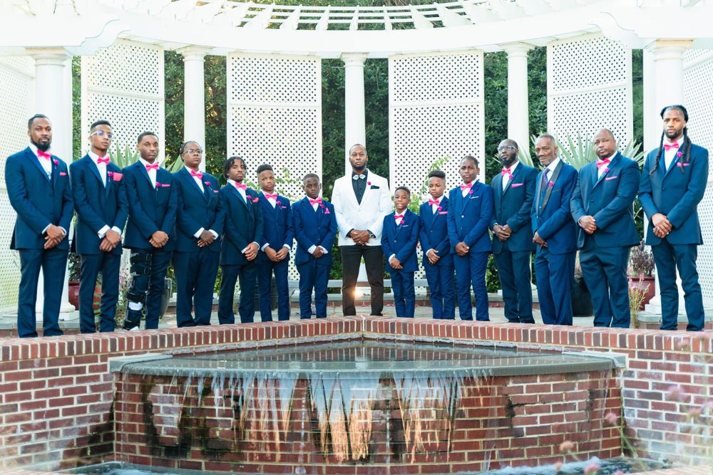 Groomsmen in navy suits pose with couple by ornate white pillared courtyard.