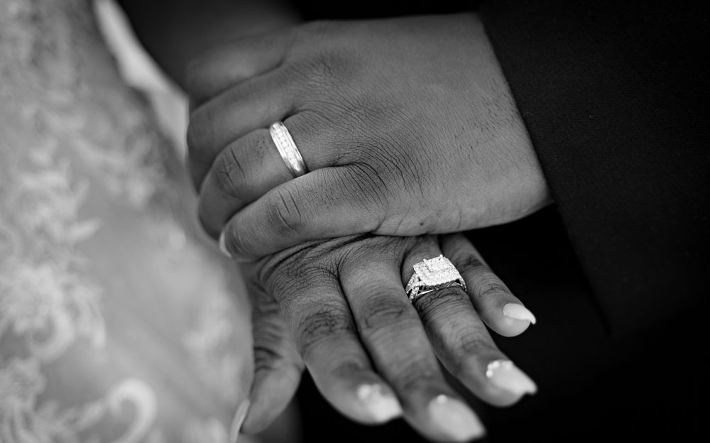 Black and white close-up of hands wearing wedding rings against lace fabric.