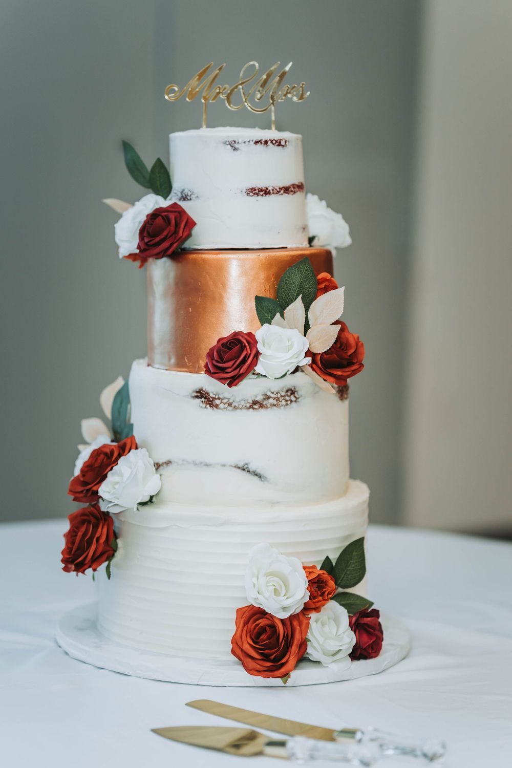 Four-tier wedding cake with white and copper metallic tiers decorated with red and white roses and a gold topper.