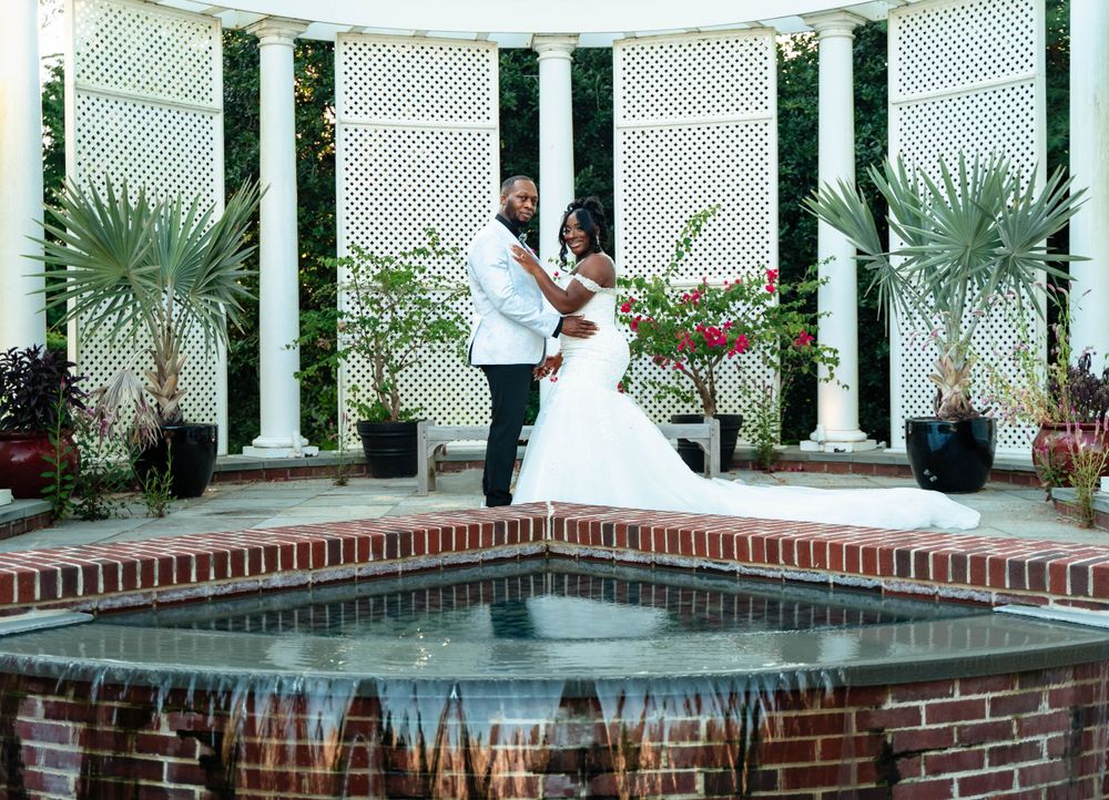 Wedding couple dancing by ornate fountain surrounded by white columns and tropical plants.
