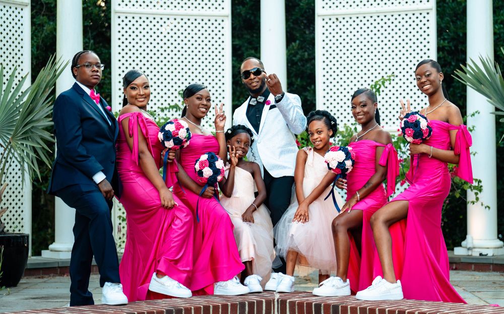 A wedding party poses together wearing vibrant pink bridesmaid dresses against a white lattice backdrop.