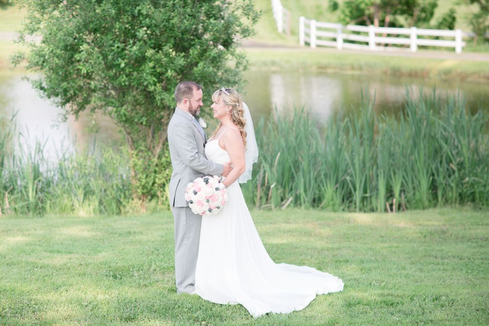 Wedding couple embracing by a scenic pond with tall grasses and white fence in background.