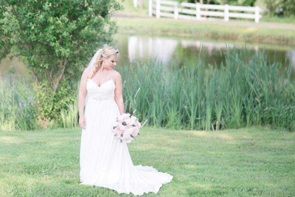 Bride in flowing white gown holding bouquet by pond with tall grasses.