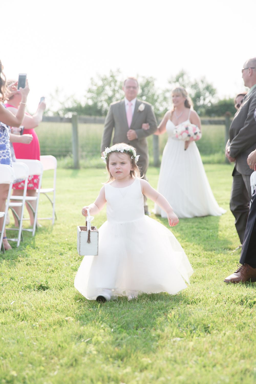 A flower girl in a white dress walks down a grassy outdoor aisle during a wedding ceremony.