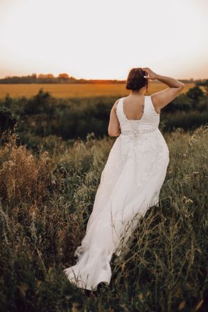 Bride in white lace wedding gown standing in golden field at sunset, viewed from behind.