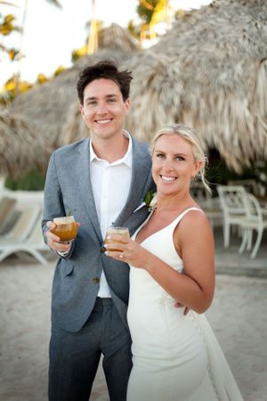 Smiling bride and groom holding tropical cocktails at beach resort wedding celebration under thatched palapa.