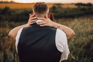 Bride embracing groom from behind with tattooed hand during a warm golden sunset in a field.