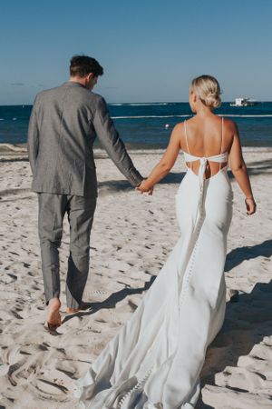 Newlywed couple holding hands walking along sandy beach, bride in backless white gown with groom in grey suit.