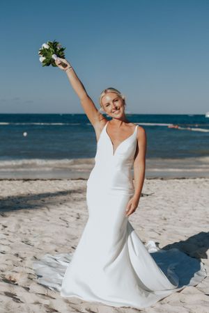 Joyful bride in white mermaid gown raising bouquet on sandy beach with blue ocean backdrop.