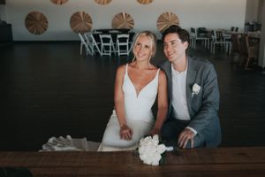 Smiling bride and groom seated together at wooden table with white bouquet in elegant wedding venue.