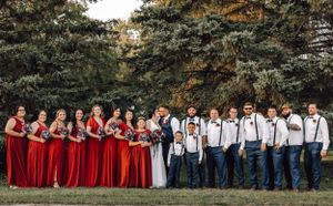 Wedding party in red bridesmaid dresses and navy suits posing outdoors under tall trees.