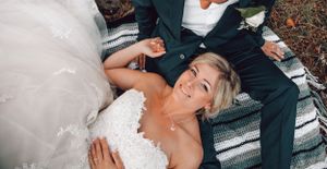 A smiling bride in a white lace wedding gown relaxes on a blanket outdoors with her groom beside her.