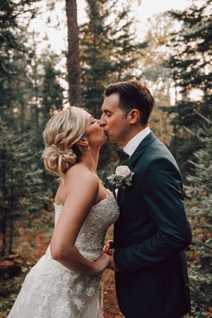 A bride and groom share a romantic kiss in a lush forest setting, with the bride in a lace gown and groom in a navy suit.