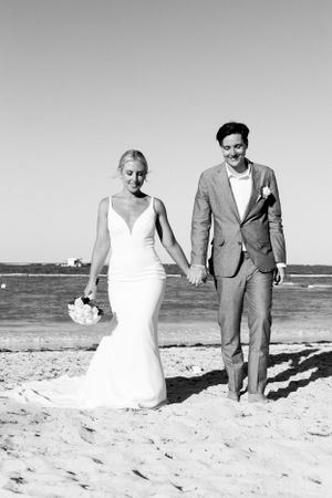 A bride and groom walk hand in hand along a sunny beach in black and white.