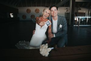 Happy newlyweds holding a Polaroid photo while seated at table with white bouquet at their wedding reception.
