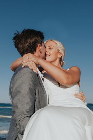 Groom lifting bride in white dress as they share a romantic kiss by the ocean on their wedding day.
