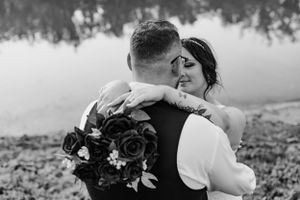 Bride and groom embracing near a reflective lake with bride holding dark roses in black and white.