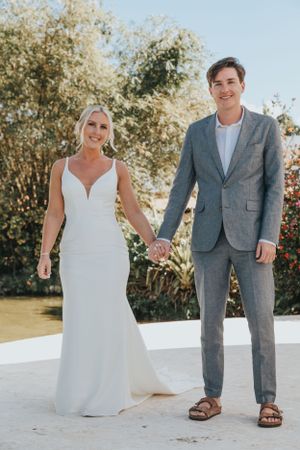 Bride in white mermaid gown and groom in grey suit holding hands outdoors with lush garden backdrop.