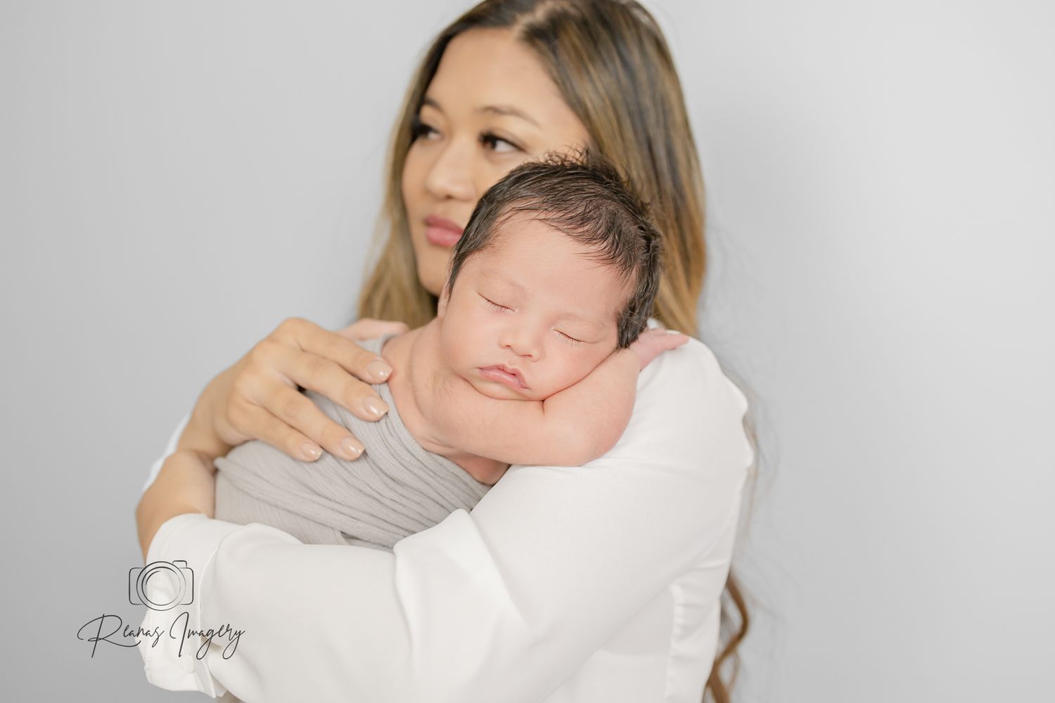 Mother gently holding her sleeping newborn baby against her shoulder in white outfit.