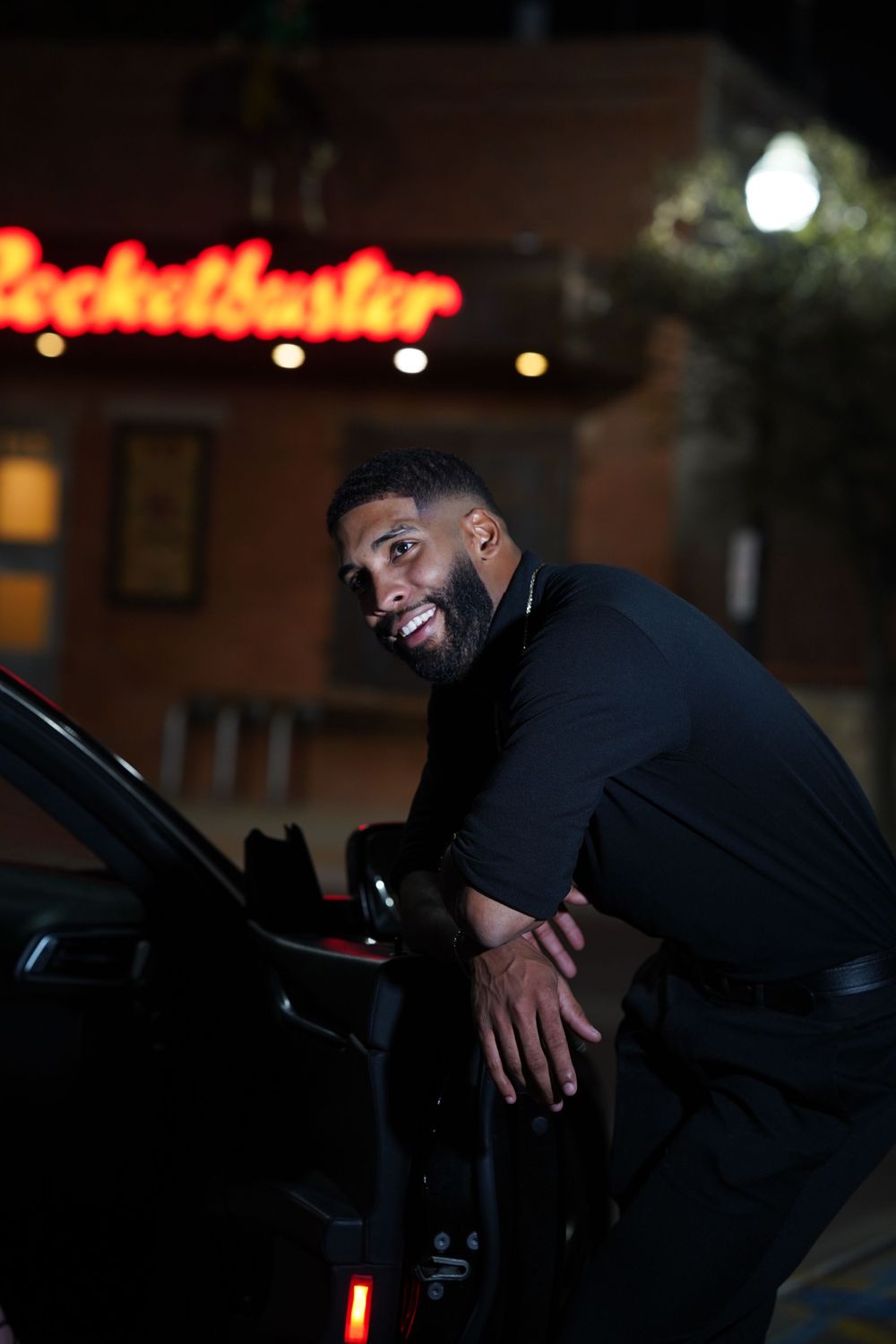 Person in dark clothing poses by car at night with neon signage visible.
