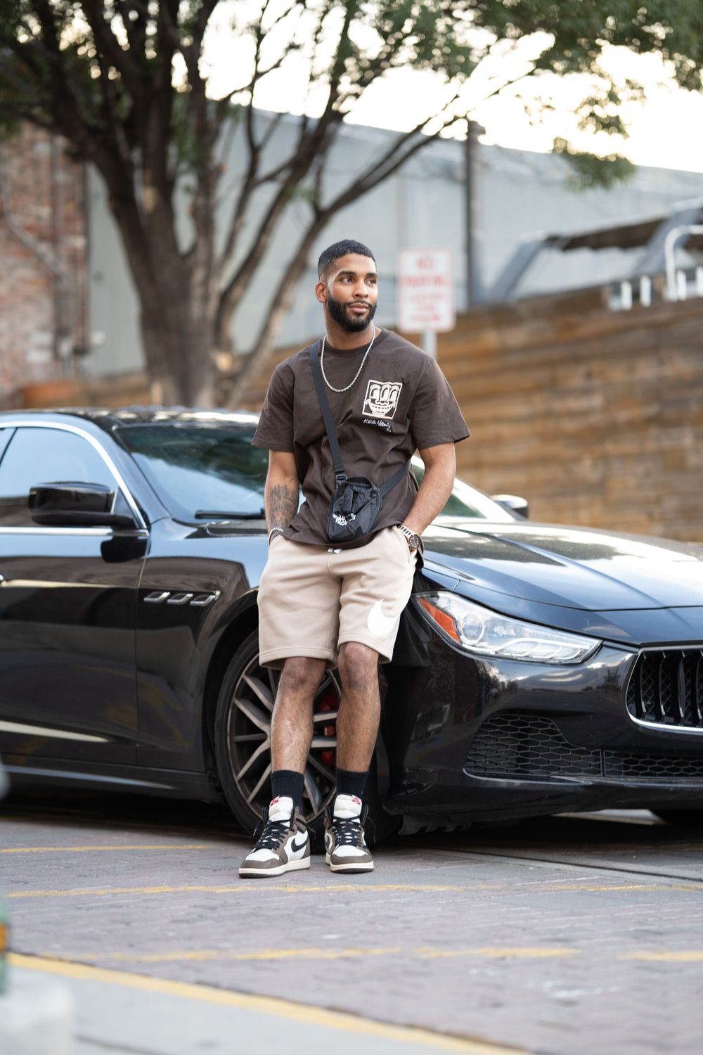 Person leaning against black Maserati wearing dark t-shirt and khaki shorts.