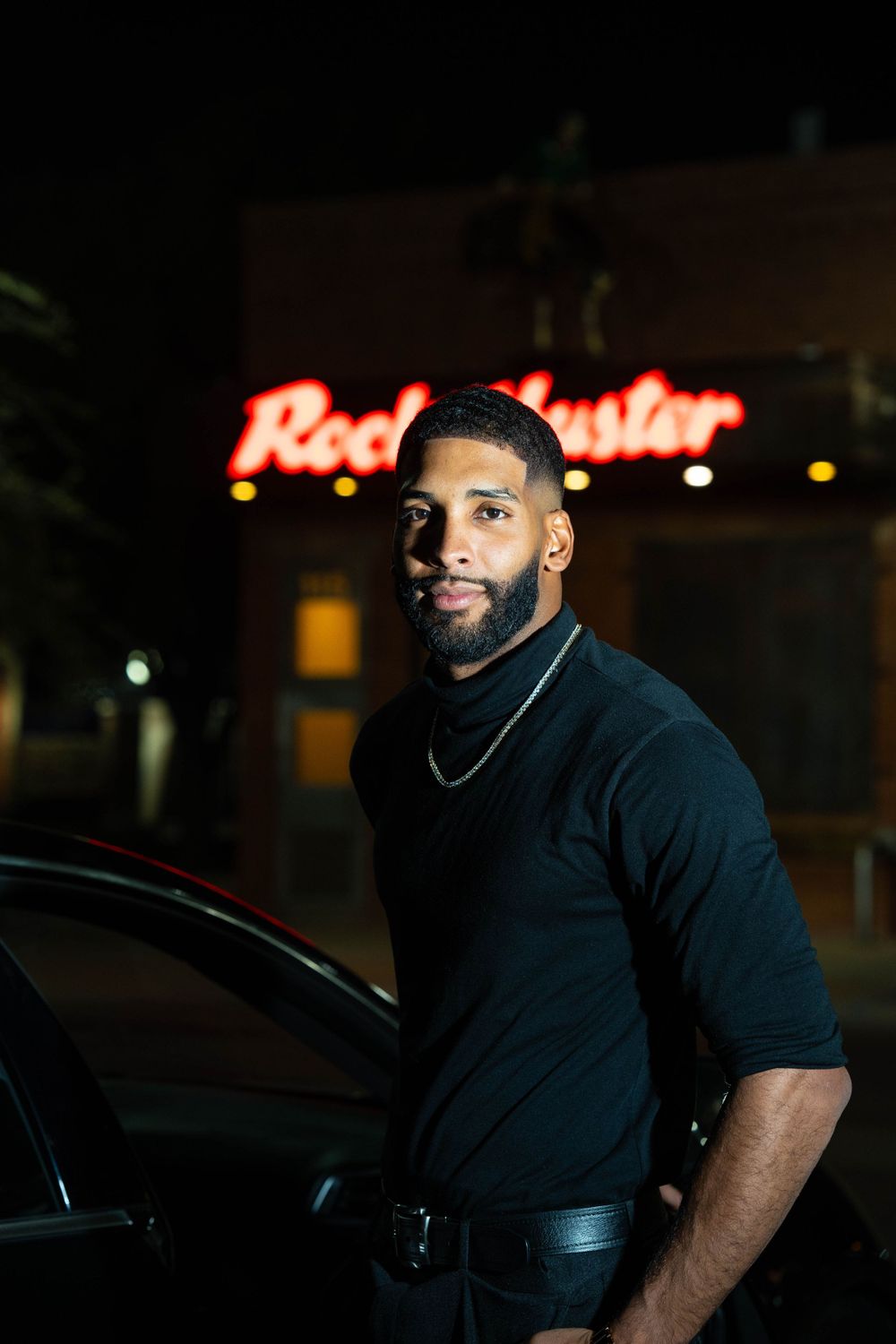 Man in black clothing leaning against car at night with illuminated signage in background.