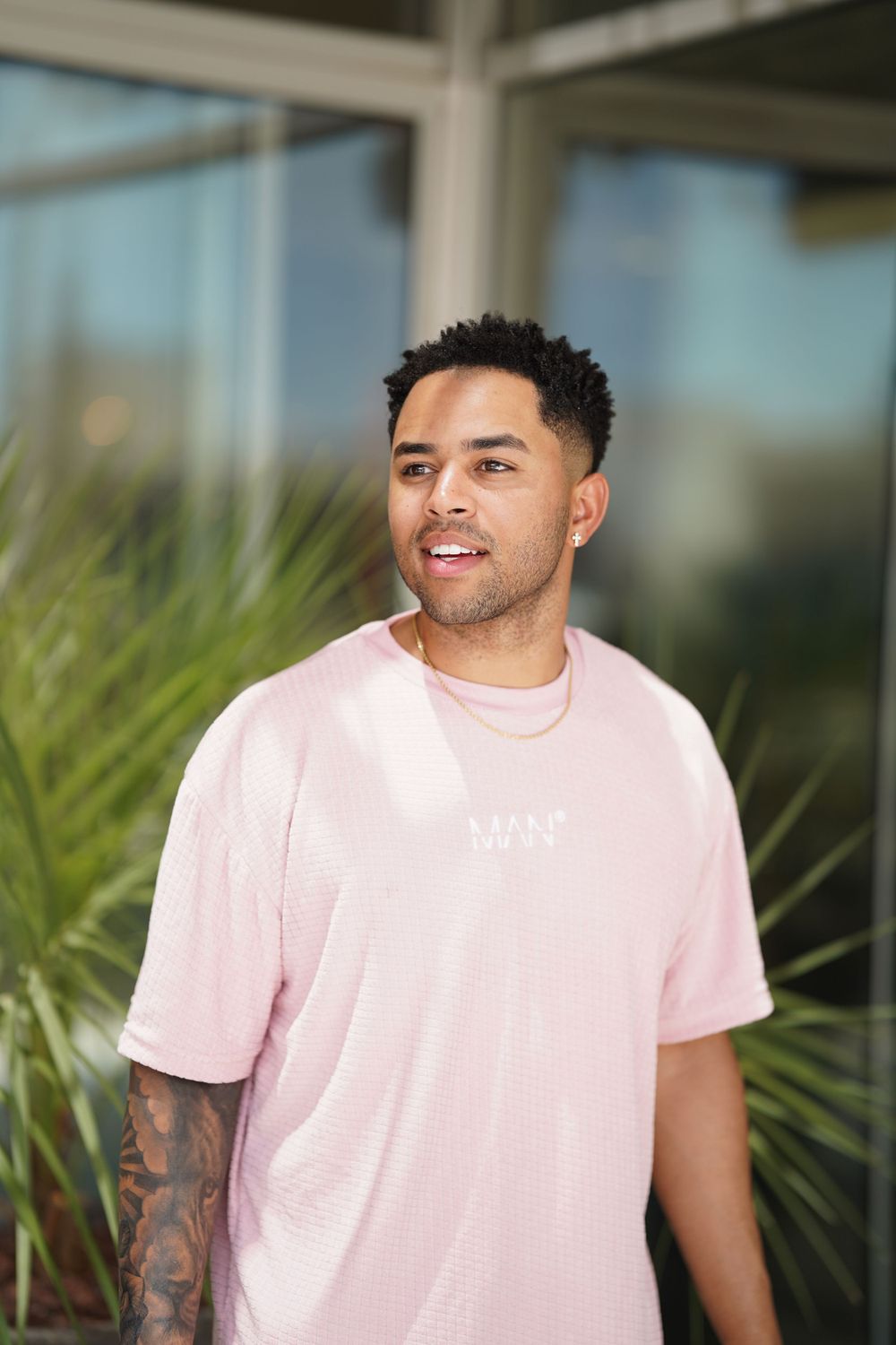 Person wearing pink t-shirt smiling outdoors near tropical plants.