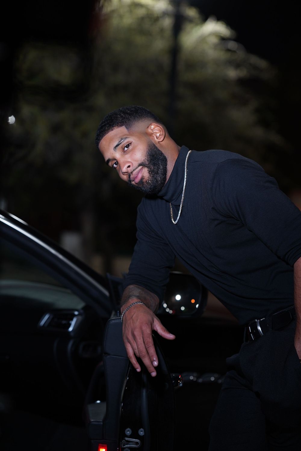 Person in black clothing posing next to car at night in moody photoshoot.