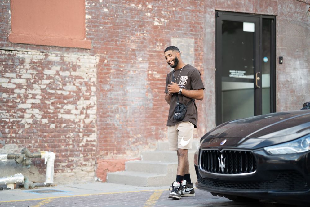 Person standing by brick wall next to black luxury car in casual streetwear.