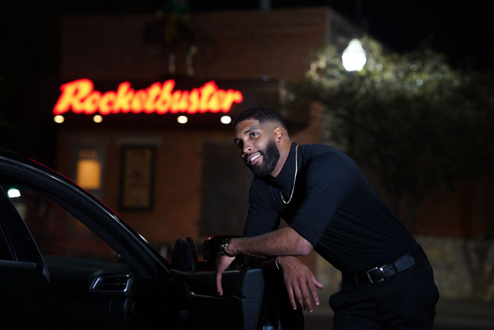 Person leaning against car at night with illuminated Blockbuster sign in background.