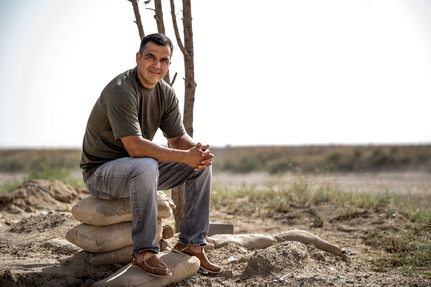 Smiling man sitting on stacked rocks in an open field with bare trees in the background.