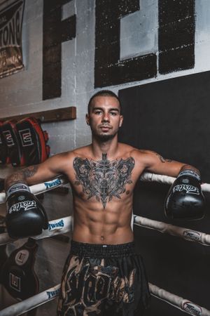 Athletic fighter with chest tattoos posing against boxing ring ropes in combat gear.