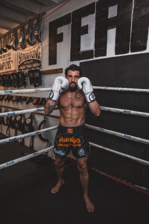 Muscular fighter in black shorts leaning against boxing ring ropes during training.