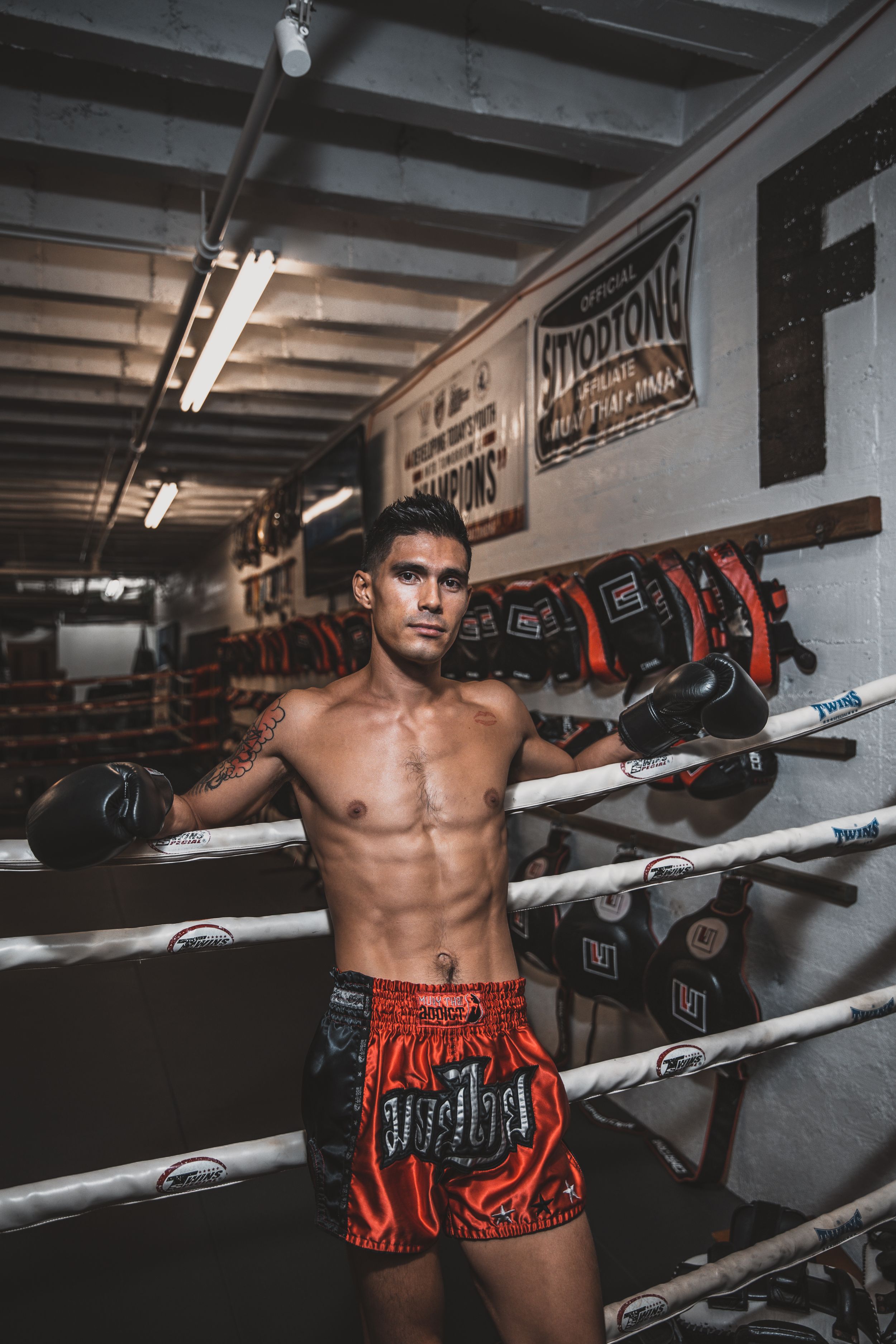Athletic boxer poses in training gym wearing red Muay Thai shorts.