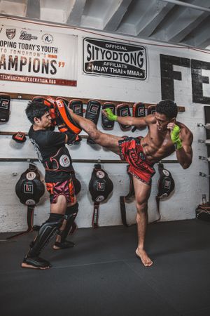 Multiple martial artists practicing low stance techniques against a wall lined with boxing equipment in a gym.