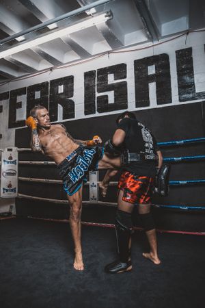 Two fighters practicing high kicks and sparring techniques in a dimly lit boxing ring.