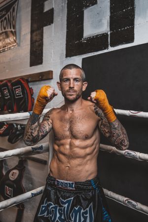Tattooed fighter with orange gloves posing in boxing stance against gym backdrop.