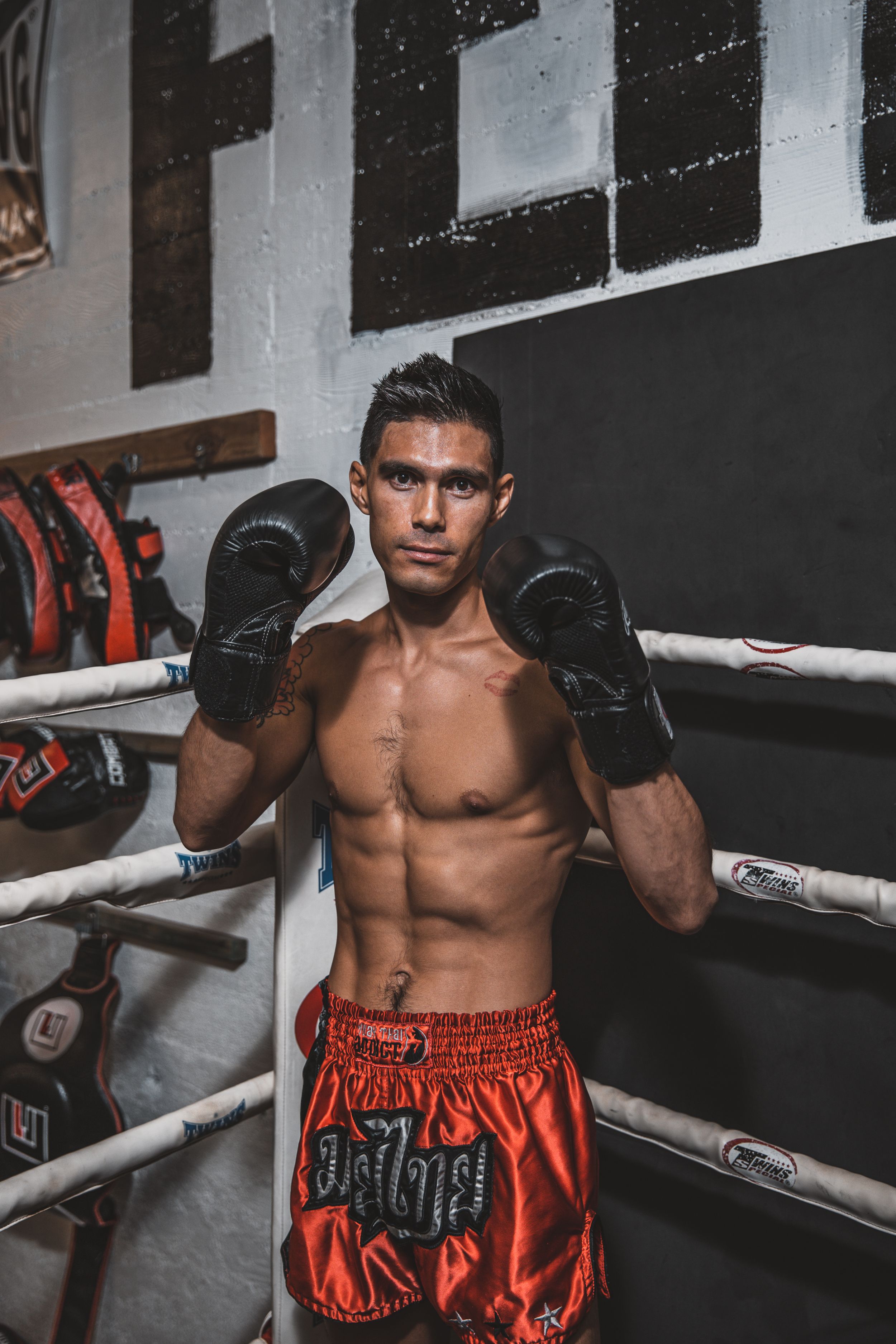 Muscular fighter in red shorts stands ready in boxing gym environment.