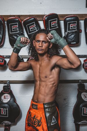 Fighter in orange shorts posing in front of wall mounted boxing equipment during training session.