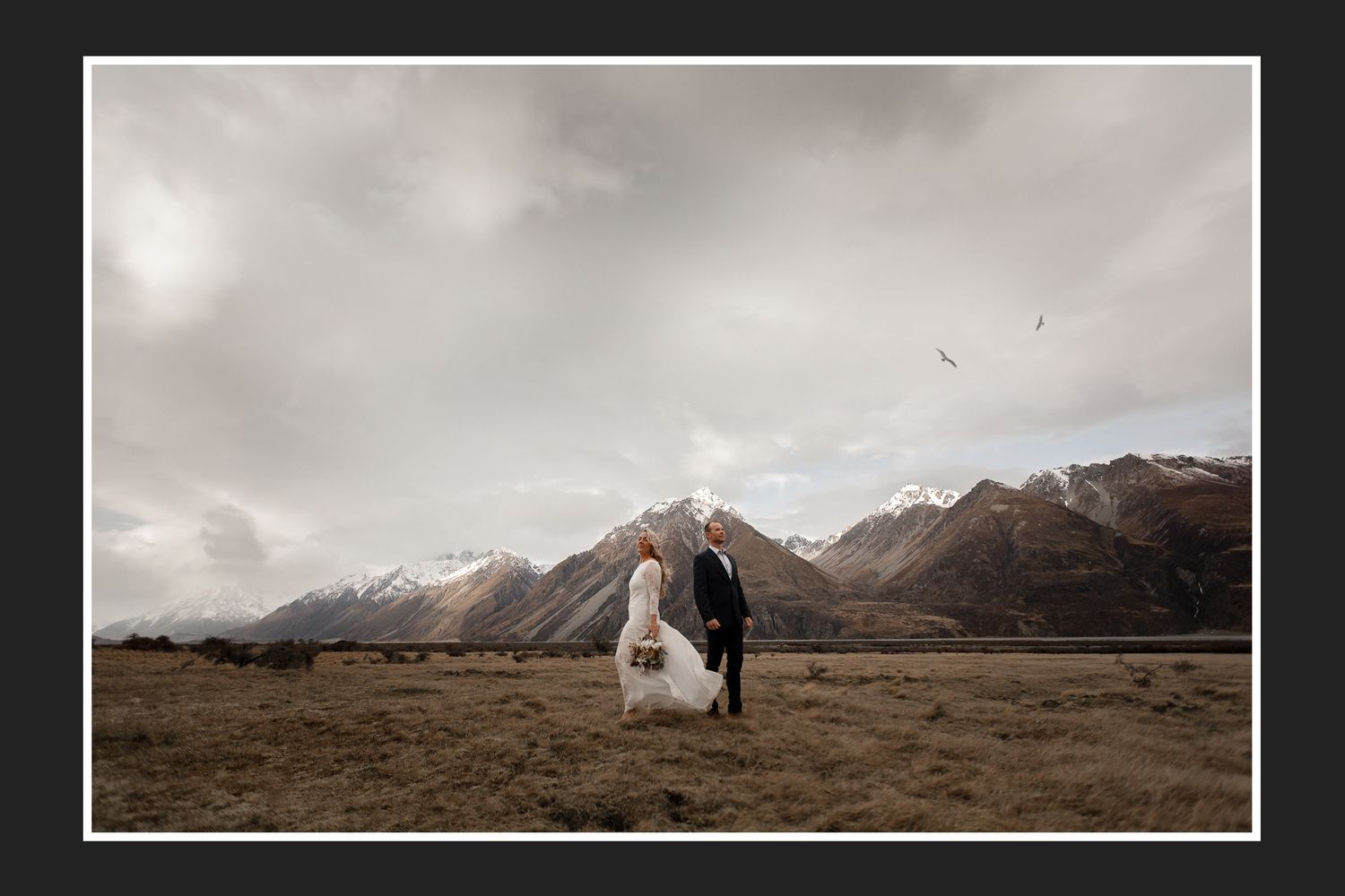 A newlywed couple stands in a vast mountain landscape with dramatic cloudy skies.
