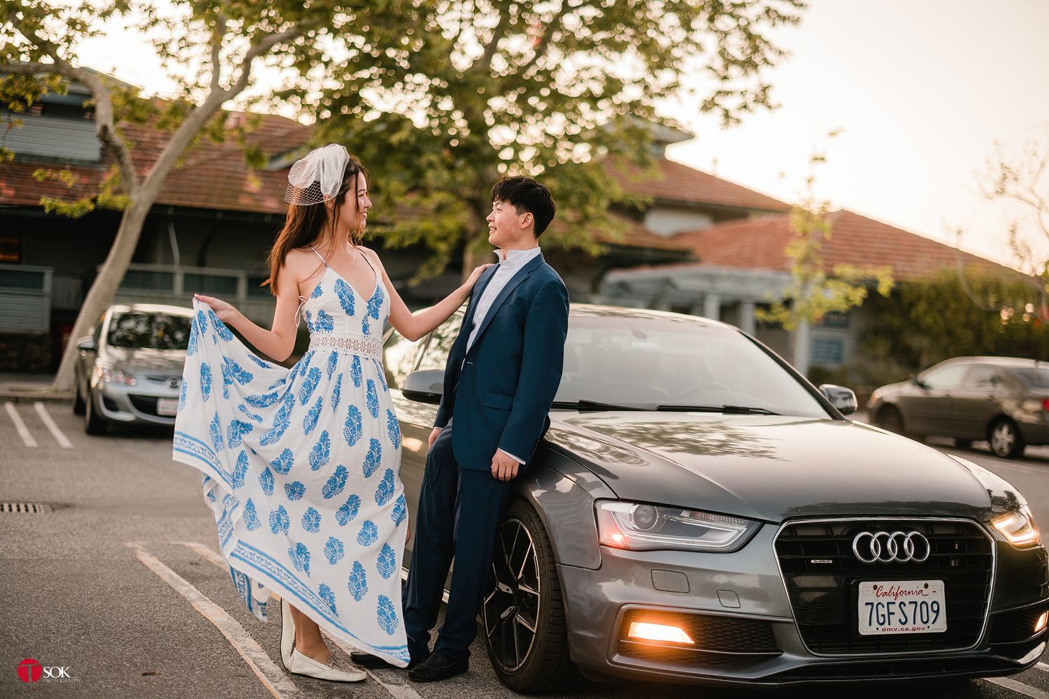 A stylish couple poses beside a silver Audi car at sunset, with her wearing a flowing blue and white dress.